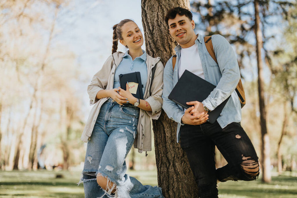 Dos jóvenes estudiantes o amigos relajándose al aire libre apoyados en un árbol con sus materiales de estudio. - Foto, imagen