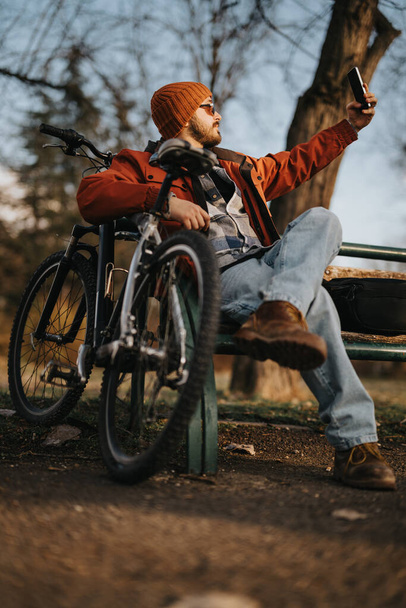 Junger Mann genießt einen gemütlichen Tag im Park mit seinem Fahrrad, surft auf seinem Handy, während er auf einer Bank sitzt. - Foto, Bild