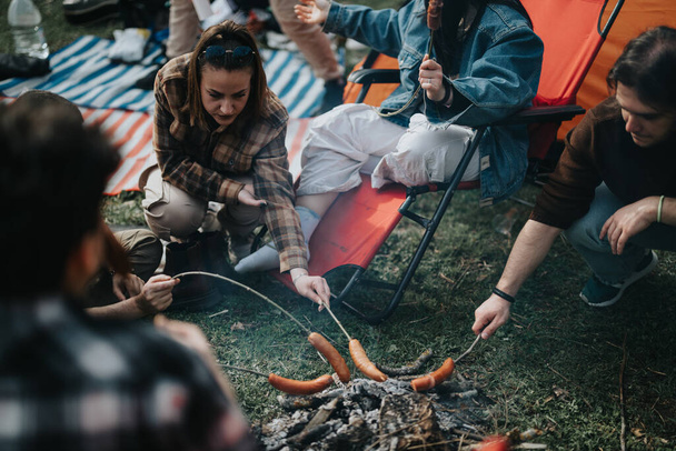キャンプファイヤーの周りに集まった友人のグループ,ソーセージを焙煎し,レイドバックの屋外キャンプ体験を楽しむ. - 写真・画像