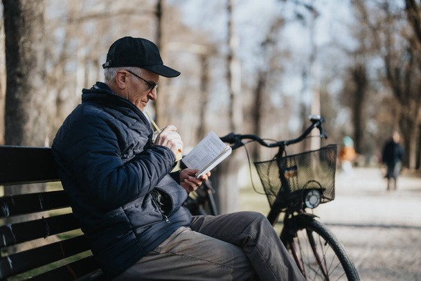 Älterer Mann genießt einen ruhigen Moment beim Lesen eines Buches auf einer Parkbank mit Fahrrad. - Foto, Bild