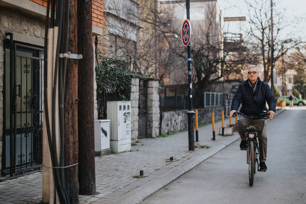 Egy idősebb úriember egy békés utcán biciklizik, aktív életmóddal és városi kerékpározással. A mindennapi élet őszinte pillanata, az idősek mobilitása és az egészséges szokások megragadása. - Fotó, kép