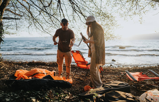 İki arkadaş kamp malzemelerini manzaralı bir göl kenarına kurup açık havanın neşesini ve rahatlığını kucaklamaktan hoşlanırlar.. - Fotoğraf, Görsel