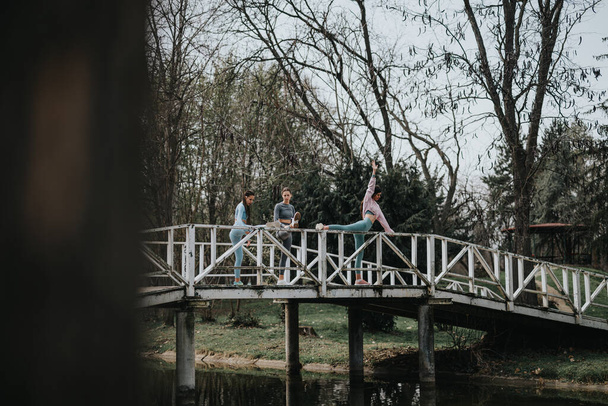 Three female athletes in workout gear stretching and exercising on a quaint bridge over a peaceful stream. - Photo, Image