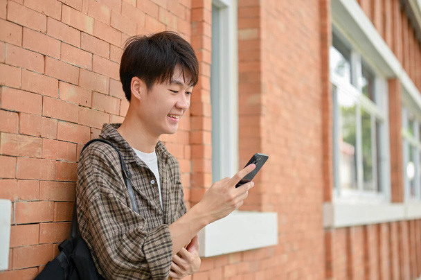 Un joven estudiante universitario asiático feliz se levanta contra una pared de ladrillo de un edificio del campus usando su teléfono inteligente. vida universitaria, estilo de vida, tecnología inalámbrica - Foto, imagen