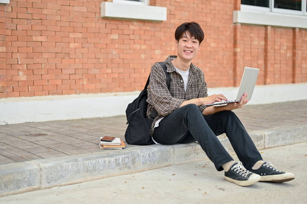 Un joven estudiante universitario asiático positivo y feliz con una mochila se sienta en la calle cerca del edificio del campus usando su computadora portátil. vida universitaria, estilo de vida, tecnología inalámbrica - Foto, imagen