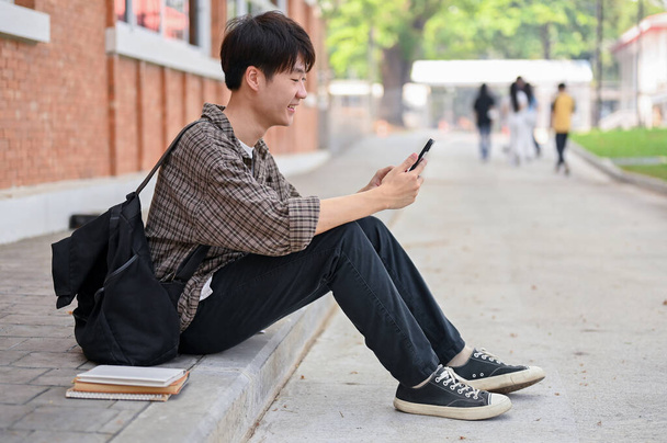A positive, happy young Asian male college student with a backpack sits on the street near the campus building using his smartphone. university life, lifestyle, wireless technology - Photo, Image