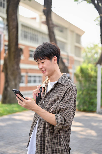 Un joven estudiante universitario asiático sonriente y feliz con ropa casual y una mochila está usando su teléfono inteligente mientras camina por el parque del campus. vida universitaria, estilo de vida, tecnología inalámbrica - Foto, imagen