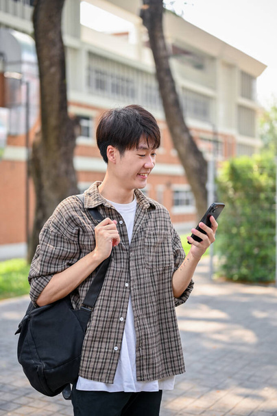 Un joven estudiante universitario asiático sonriente y feliz con ropa casual y una mochila está usando su teléfono inteligente mientras camina por el parque del campus. vida universitaria, estilo de vida, tecnología inalámbrica - Foto, imagen