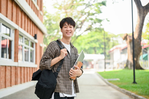 Un joven estudiante universitario asiático alegre en ropa casual, llevando una mochila, se encuentra fuera de un edificio de ladrillo en el campus, sosteniendo un ordenador portátil y libros, sonriendo a la cámara. - Foto, imagen