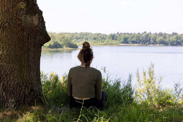 Cette image évocatrice représente une femme par derrière, assise dans l'immobilité au bord d'un lac tranquille. Flanquée d'un tronc d'arbre robuste et surplombant les eaux calmes, sa posture suggère une relaxation profonde ou - Photo, image