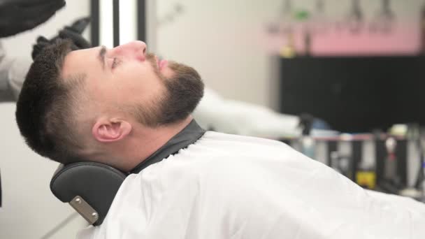 Barber brengt een zwart hydraterende masker op een mans gezicht tijdens het scheren. Een dermatoloog brengt een zwart masker aan op het gezicht van een bebaarde patiënt om poriën en rimpels te verwijderen 4k video. - Video