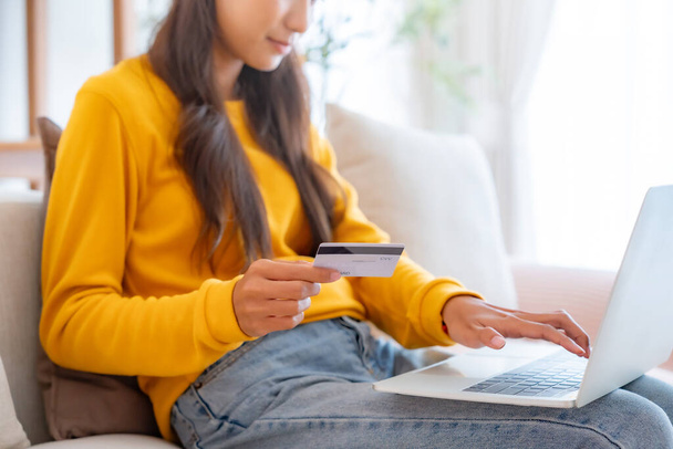 Fiatal ázsiai nő ül a kanapén a nappaliban, online banki fizetéseket bonyolít az interneten keresztül bankkártyáról számítógépes laptopon. Online vásárlás a notebookon hitelkártyával - Fotó, kép