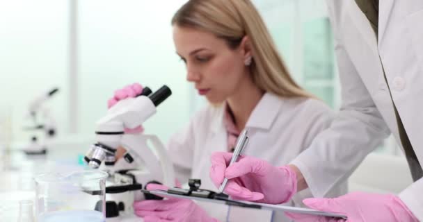 scienziato femminile utilizza un microscopio e prende appunti. Lavoro collaborativo di assistenti di laboratorio in un laboratorio - Filmati, video