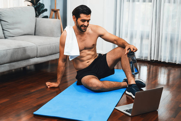 Sportos és sportos férfi pihen fitnesz szőnyegen az online testedzés edzés során a fitt fizikum és az egészséges sport életmód otthon. Vidám otthoni edzés képzési koncepció. - Fotó, kép