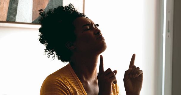 Dindar genç siyah bir kadın sessizce evde dua ediyor Tanrı 'dan yardım ve destek arıyor gözleri kapalı ve gökyüzünü işaret ediyor inanç ve ruhani duygular içinde. - Fotoğraf, Görsel