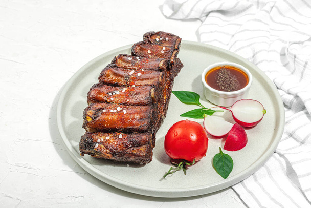 Baked pork ribs with spices, vegetables and sauce. Traditional ingredient for BBQ, picnic concept. Composition with glazed meat, linen napkin, trendy plate. Hard light, dark shadow, copy space - Photo, Image
