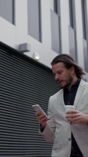 Joven hombre caucásico charlando trabajando en su teléfono inteligente. Empresario escribiendo mensaje y caminando cerca del edificio industrial. Ejecutivo bebiendo café para ir durante su descanso - Metraje, vídeo