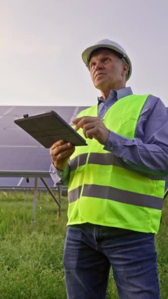 A mérnök a napelemek előtt áll. Tabletta tartása és a fotovoltaikus napelemek ellenőrzése. 4K - Felvétel, videó