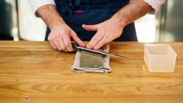 fechar um chef em uma jaqueta branca na cozinha na mesa afia uma faca em um fogão verde - Filmagem, Vídeo