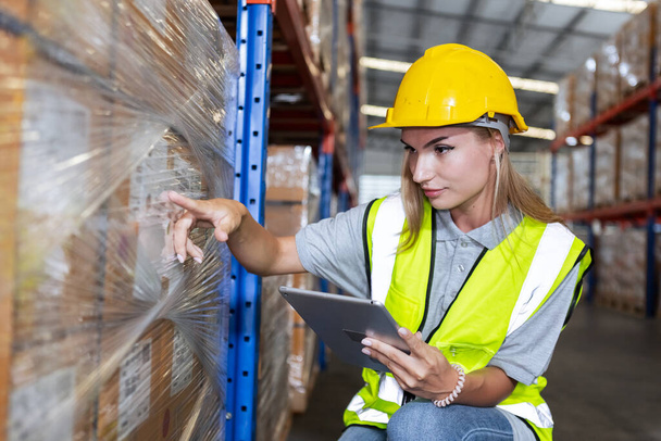 Vrouwelijke werknemer draagt veiligheidsuniform en harde hoed met behulp van tabletcontrole inspecteren goederen op rekken in magazijn. vrouwen werknemer controleren voorraad inspecteren product in fabriek. - Foto, afbeelding