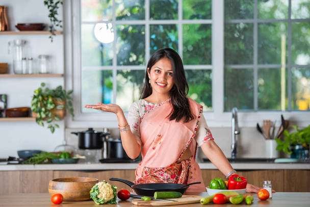 Hintli genç bir kadın mutfak aletlerini, tencereyi, spatulayı, boş tabağı işaret ediyor, itiyor ya da teşvik ediyor. - Fotoğraf, Görsel