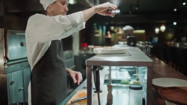 Professionele mannelijke chef-kok in schort en hoed zout eten geserveerd op bord tijdens het bereiden van orde in de open keuken in restaurant - Video
