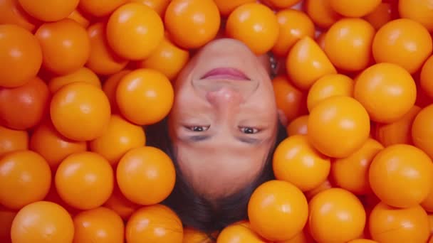 Πρόσωπο βυθισμένο σε μια θάλασσα από λαμπερές πορτοκαλί μπάλες, δείχνοντας μόνο το χαμογελαστό πρόσωπό τους κατά τη διάρκεια του πρωινού - Πλάνα, βίντεο