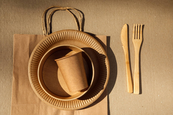 Комплект бумажной посуды и деревянных столовых приборов на коричневом фоне с бумажным пакетом. Экологически чистые, нулевые отходы. Вид сверху - Фото, изображение