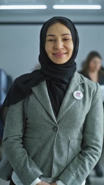 La mujer se encuentra en un moderno centro de votación, posa, sonríe y mira a la cámara. Retrato de la mujer árabe, votante de las elecciones de los Estados Unidos de América. Antecedentes con mesas de votación. Concepto de deber cívico. - Imágenes, Vídeo