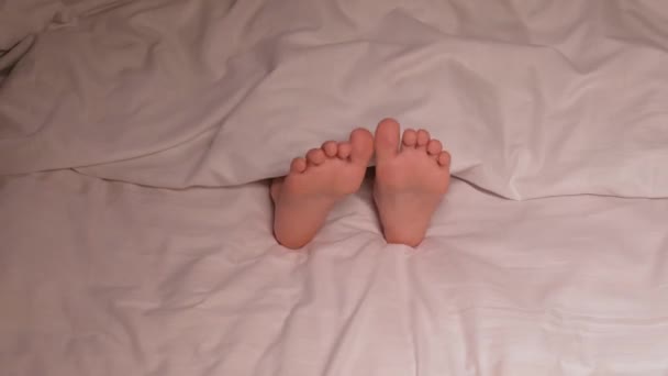 Dziecięce stopy pod kocem na prześcieradle wieczorem. Śpiąca dziewczynka z gołymi nogami kręci nogami we śnie na wygodnym łóżku w sypialni - Materiał filmowy, wideo