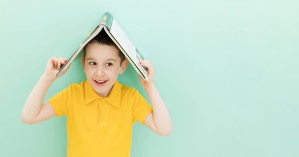 Niño de escuela caucásico sostiene libro en una cabeza sobre un fondo verde claro con espacio de copia - Foto, Imagen