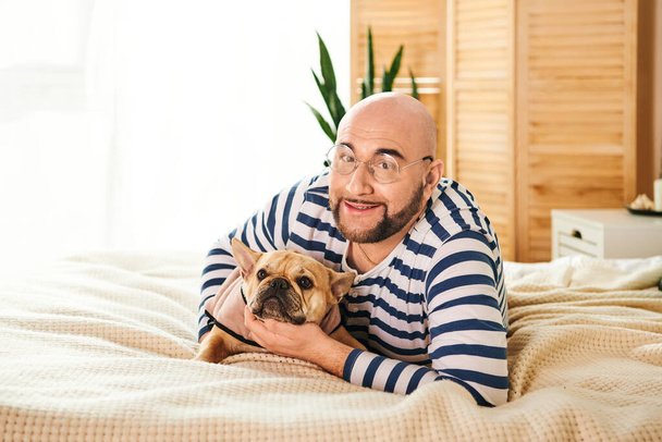 Ein Mann umarmt friedlich eine kleine französische Bulldogge, während er auf einem Bett liegt. - Foto, Bild
