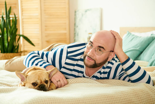 Silmälasipäinen mies makaa rauhallisesti sängyllä ranskalaisen bulldoginsa kanssa.. - Valokuva, kuva