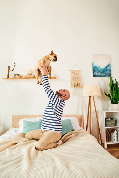 Relaxed man with glasses lovingly lifts French Bulldog in air on cozy bed. - Photo, Image