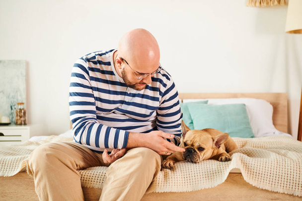 Un uomo in occhiali si siede su un letto, accarezzando affettuosamente un bulldog francese. - Foto, immagini