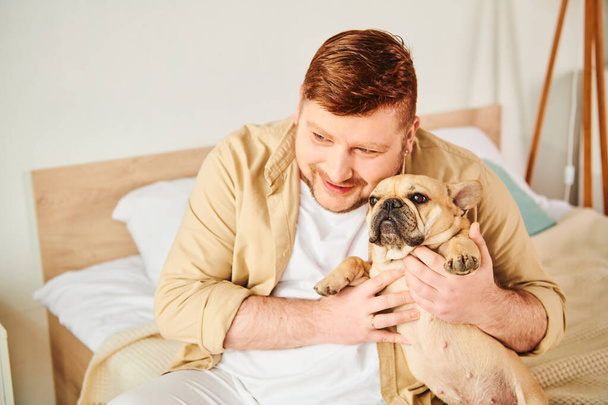 Un hombre elegante sentado en una cama, sosteniendo amorosamente un pequeño Bulldog francés. - Foto, imagen