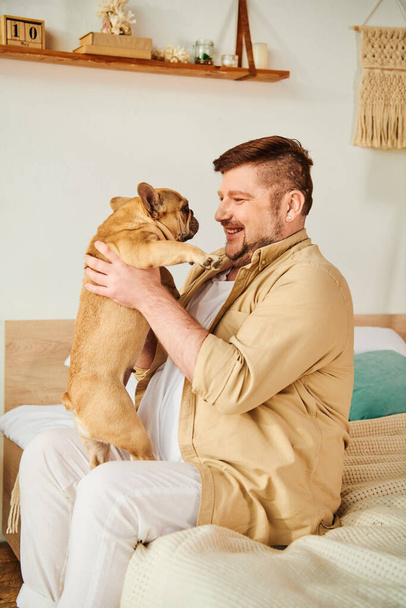 Un hombre sentado en una cama, sosteniendo amorosamente a un bulldog francés. - Foto, imagen