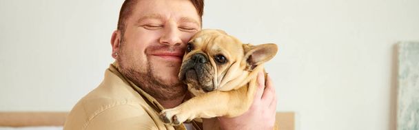 Schöner Mann, der zu Hause eine kleine französische Bulldogge in den Arm nimmt. - Foto, Bild