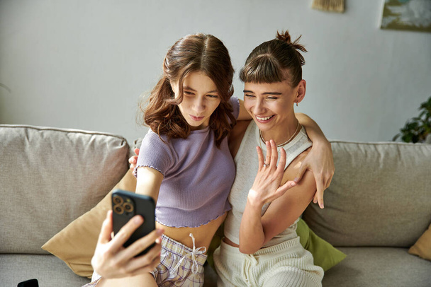 Lésbicas casal em trajes aconchegantes sentado no sofá, absorvido no telefone celular. - Foto, Imagem