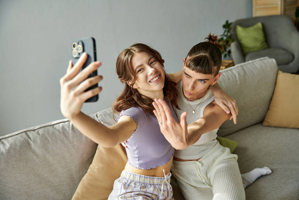 Dos mujeres en trajes cómodos sonriendo mientras se toman una selfie en un sofá. - Foto, Imagen