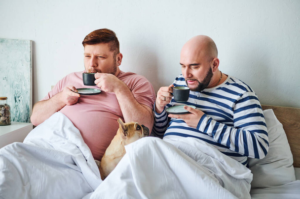 Un couple d'hommes et leur bouledogue français assis ensemble sur un lit, profitant d'un moment paisible. - Photo, image