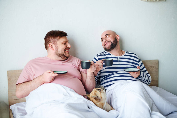 Ένα γκέι ζευγάρι χαλαρώνει στο κρεβάτι με το Γαλλικό μπουλντόγκ κοντά.. - Φωτογραφία, εικόνα