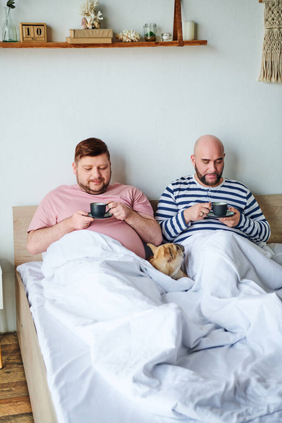 Una pareja gay se sienta junta en una cama, acompañada por su bulldog francés. - Foto, imagen