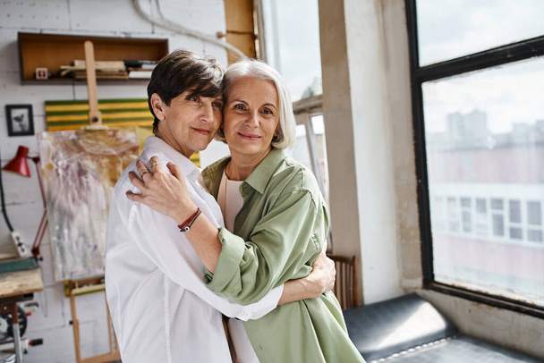 A mature lesbian couple hugging tenderly in an art studio. - Foto, imagen