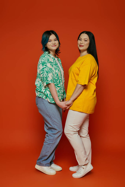 An Asian mother and her teenage daughter standing gracefully beside each other against a vibrant red background. - Zdjęcie, obraz