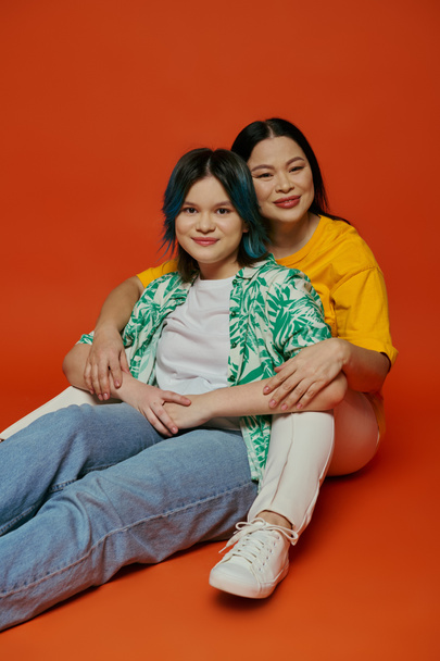 アジアの母親と10代の娘が地面に座って,オレンジ色のスタジオの背景に写真を撮ります.. - 写真・画像