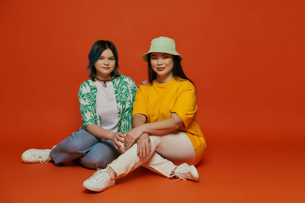 アジア人の母親と10代の娘が,オレンジ色の背景にあるスタジオで優雅にポーズをとって地面に座った.. - 写真・画像