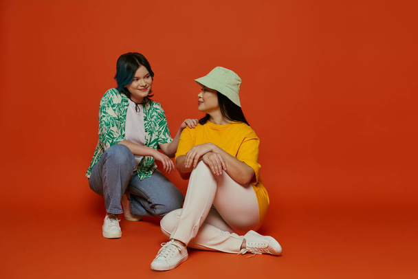 Asian mother and her teenage daughter sit closely, sharing a moment of connection, on a vibrant orange backdrop. - Foto, afbeelding
