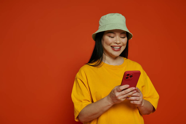 Asian woman wearing a yellow t-shirt joyfully holds a red cell phone in a studio with an orange background. - Photo, Image