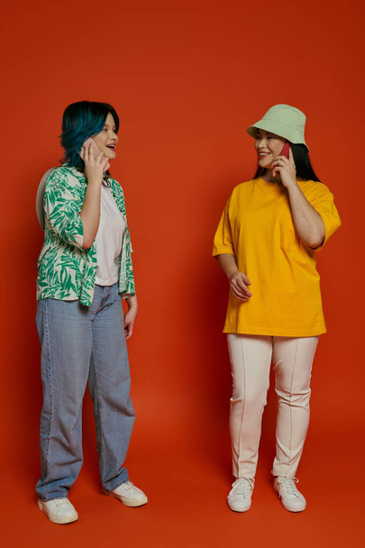 a mother and her teenage daughter, standing together on an orange background, engaged in separate phone conversations. - Photo, Image